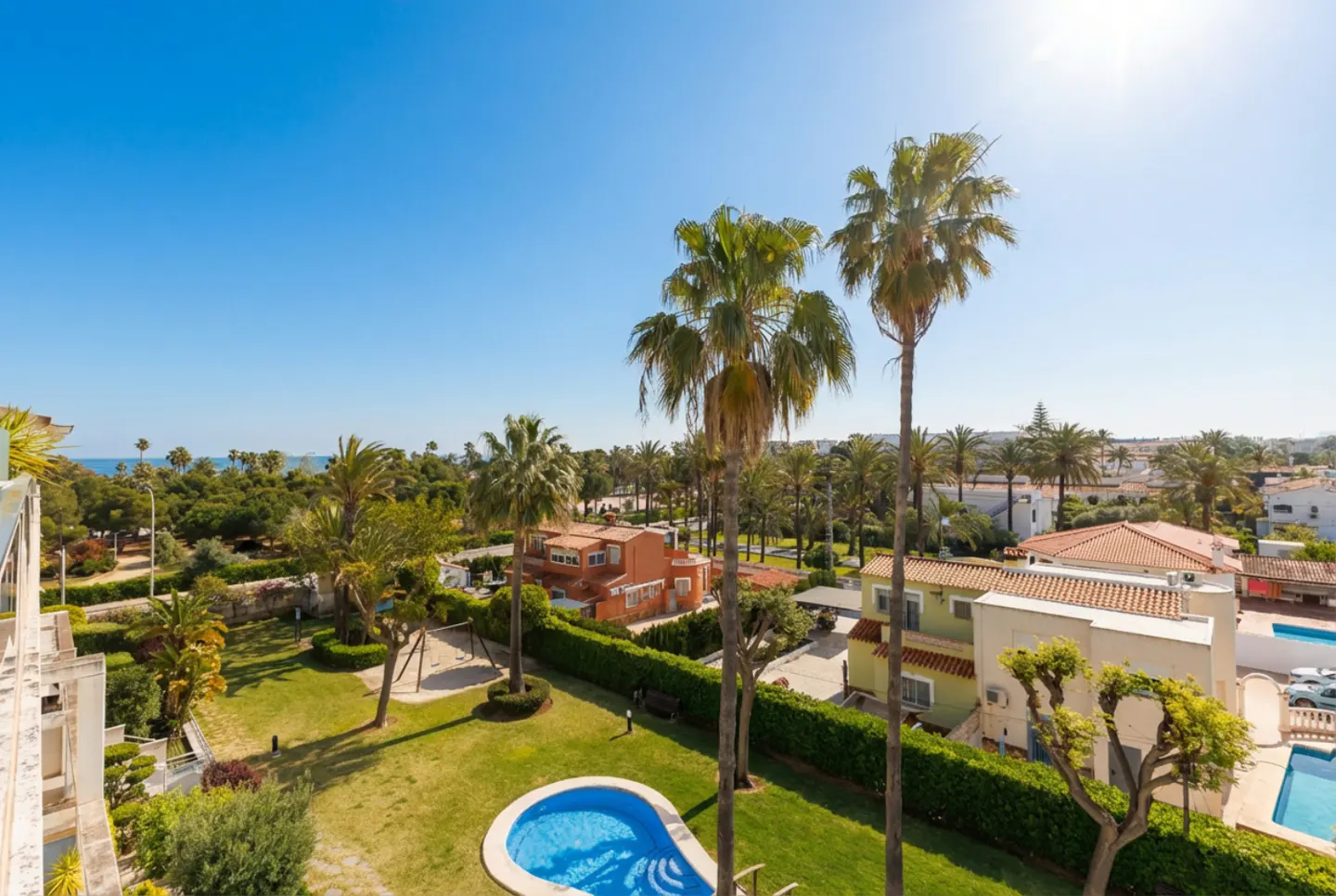 Aerial view of a property with a blue pool, green lawn, palm trees, and houses under a clear blue sky.
