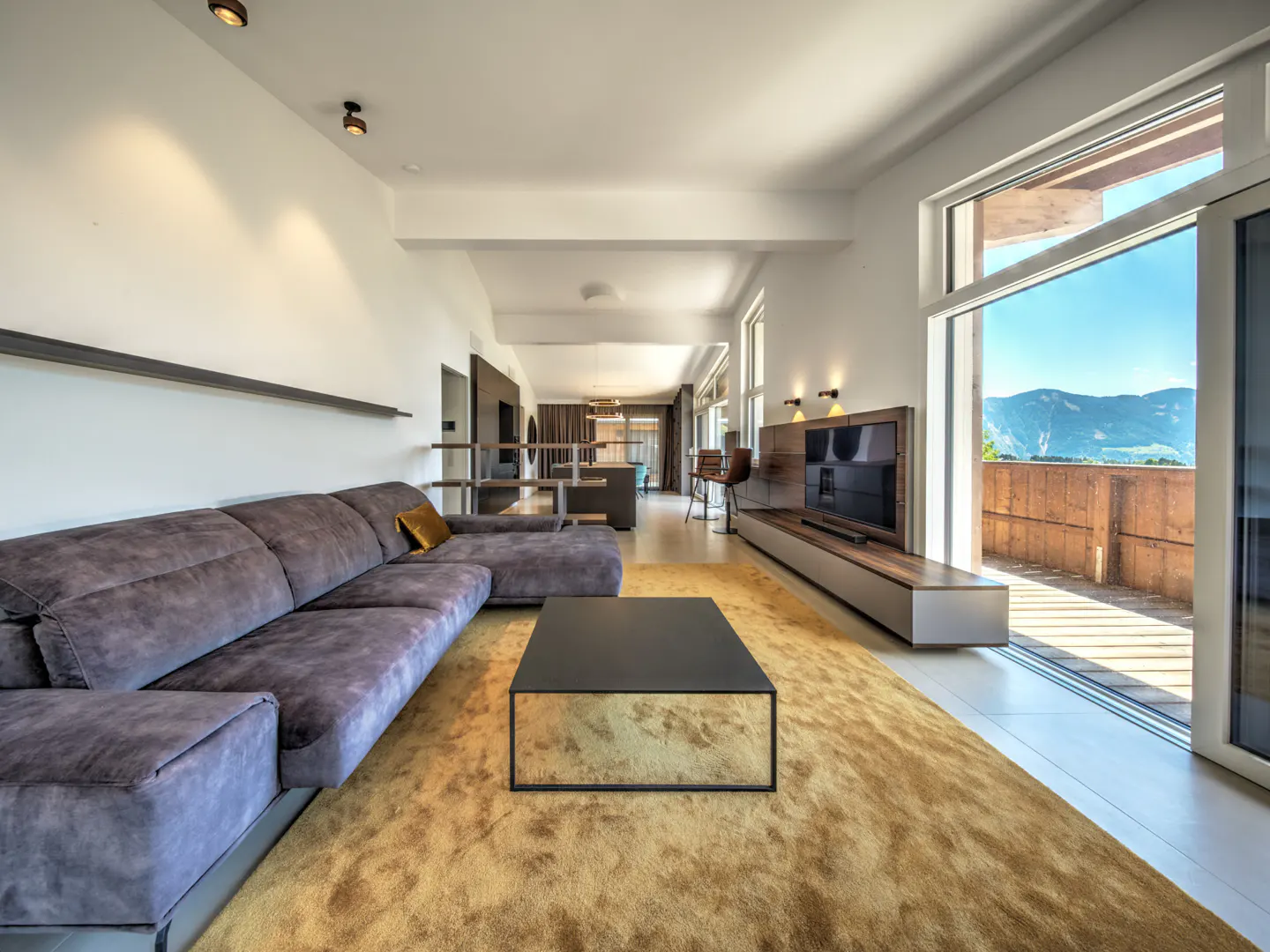Modern living room with a grey sectional sofa, black coffee table, gold rug, and a balcony view of mountains.