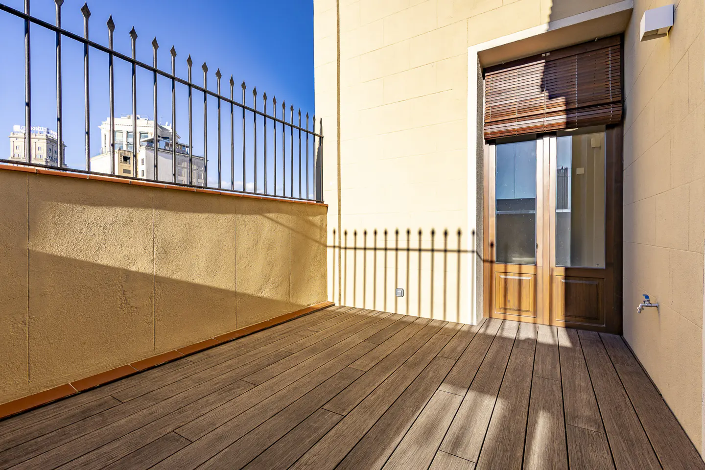 A sunlit balcony with a wooden floor, a metal railing, and a tan wall with a door. Buildings are visible in the background.