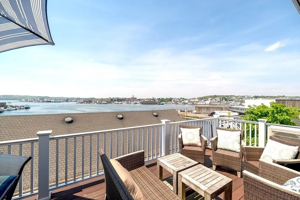 Sunny rooftop deck with wicker furniture, white railings, and a panoramic view of a harbor and coastal town.
