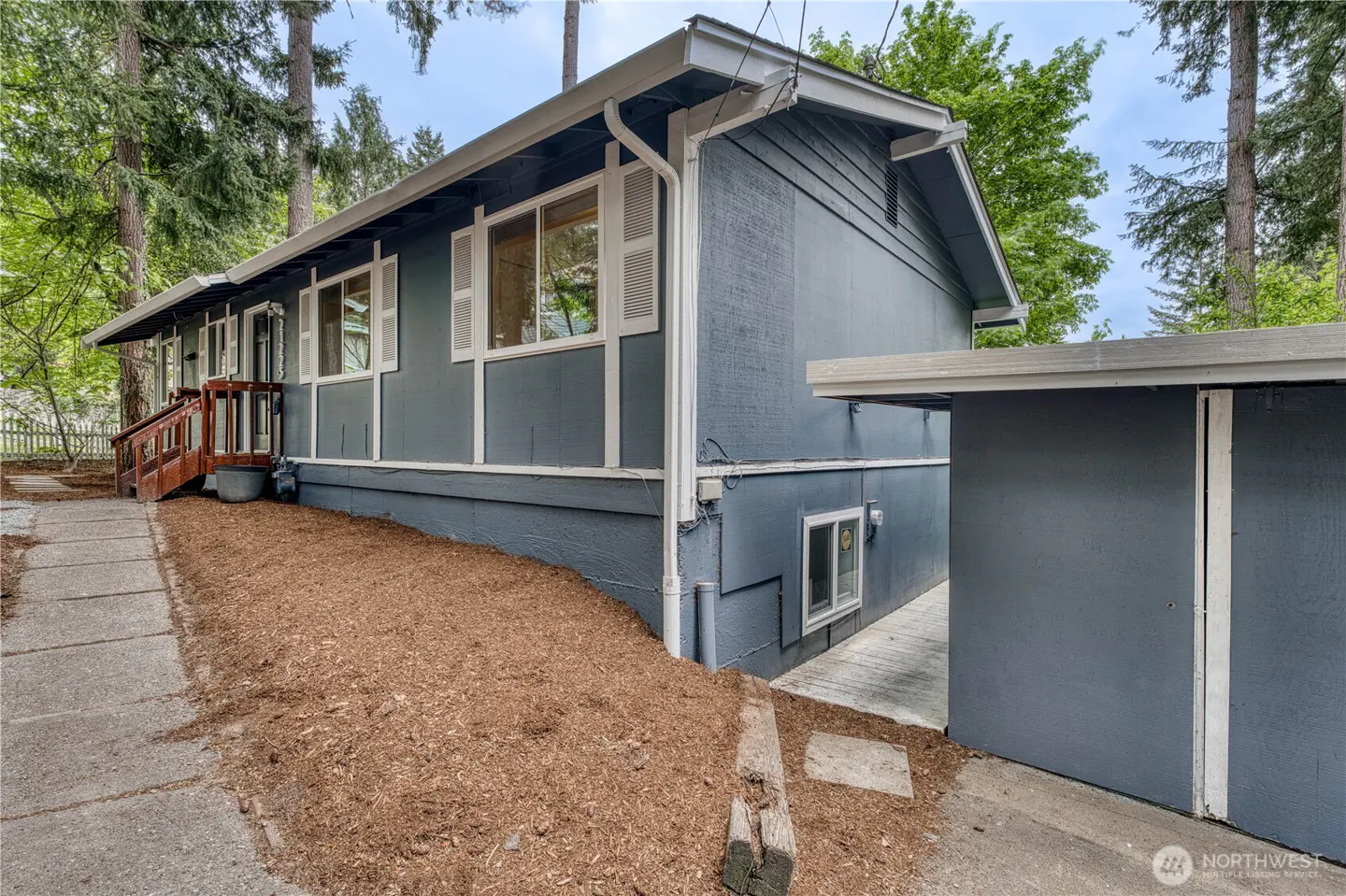 Exterior view of a gray house with white trim and shutters, surrounded by trees and mulch. A concrete walkway leads to the front porch.
