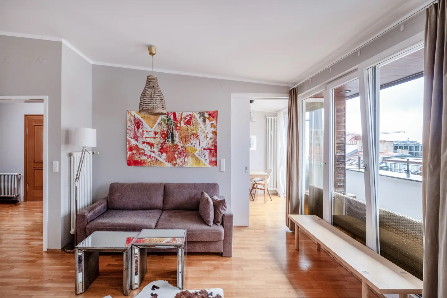 Living room with brown sofa, mirrored tables, and abstract art. Balcony access with city view. Hardwood floors and gray walls.