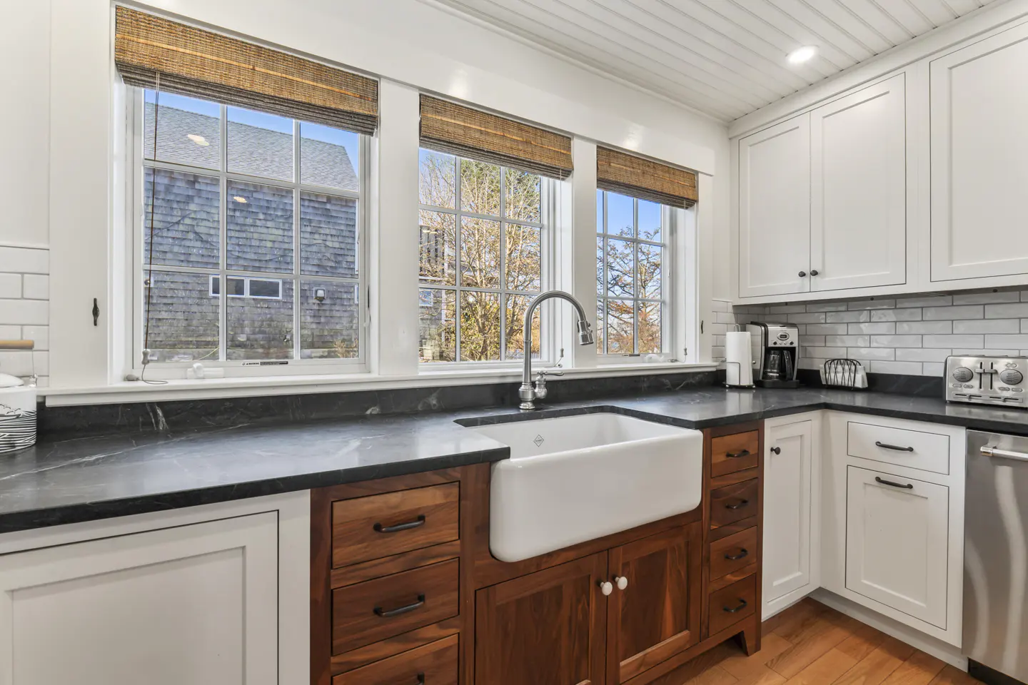 Bright kitchen with white cabinets, wood drawers, and a farmhouse sink under three windows with woven shades.