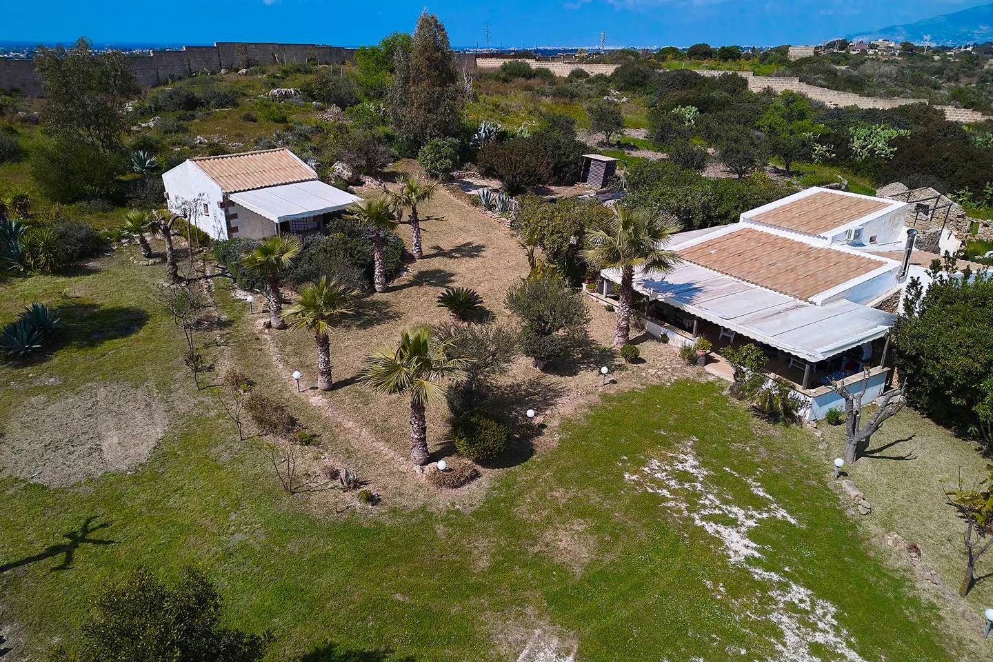 Aerial view of two white houses with terracotta roofs, surrounded by green lawns, trees, and palm trees.