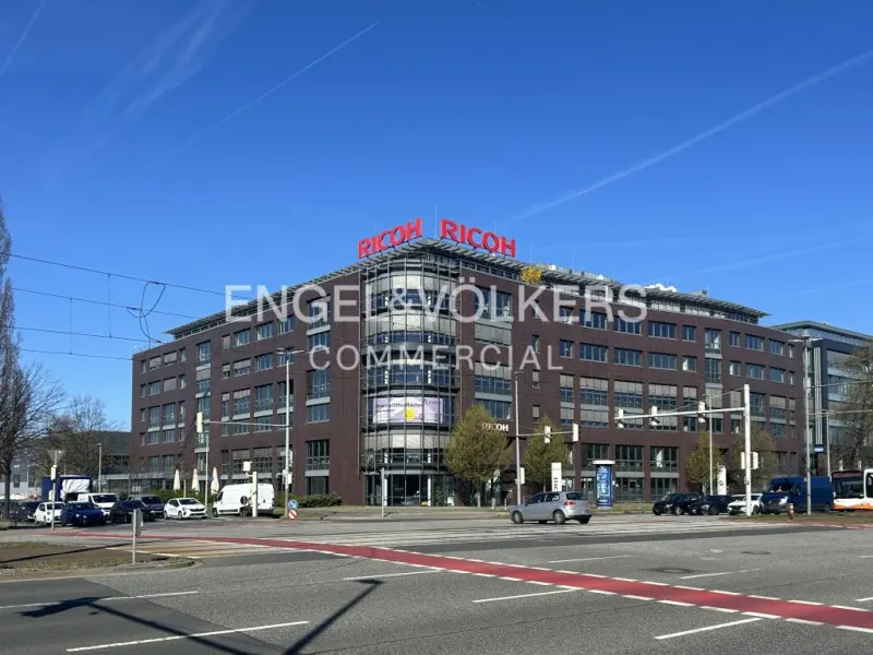Commercial building with "RICOH" sign on top, brown brick facade, and Engel & Völkers logo. Cars on street, blue sky.