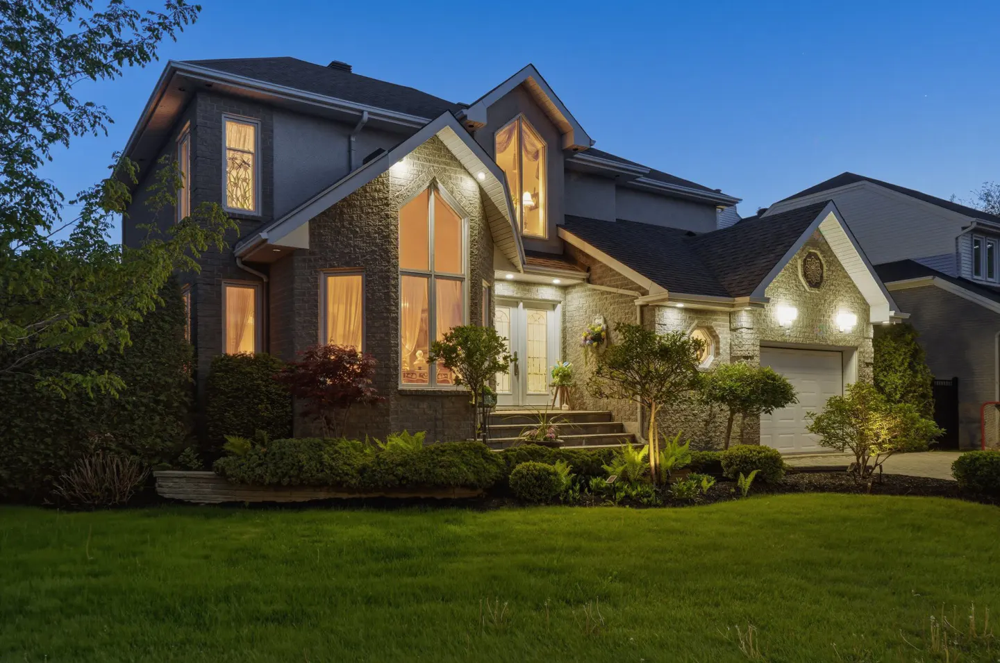 Two-story gray stone house at dusk with illuminated windows, a white garage, and a manicured front lawn.