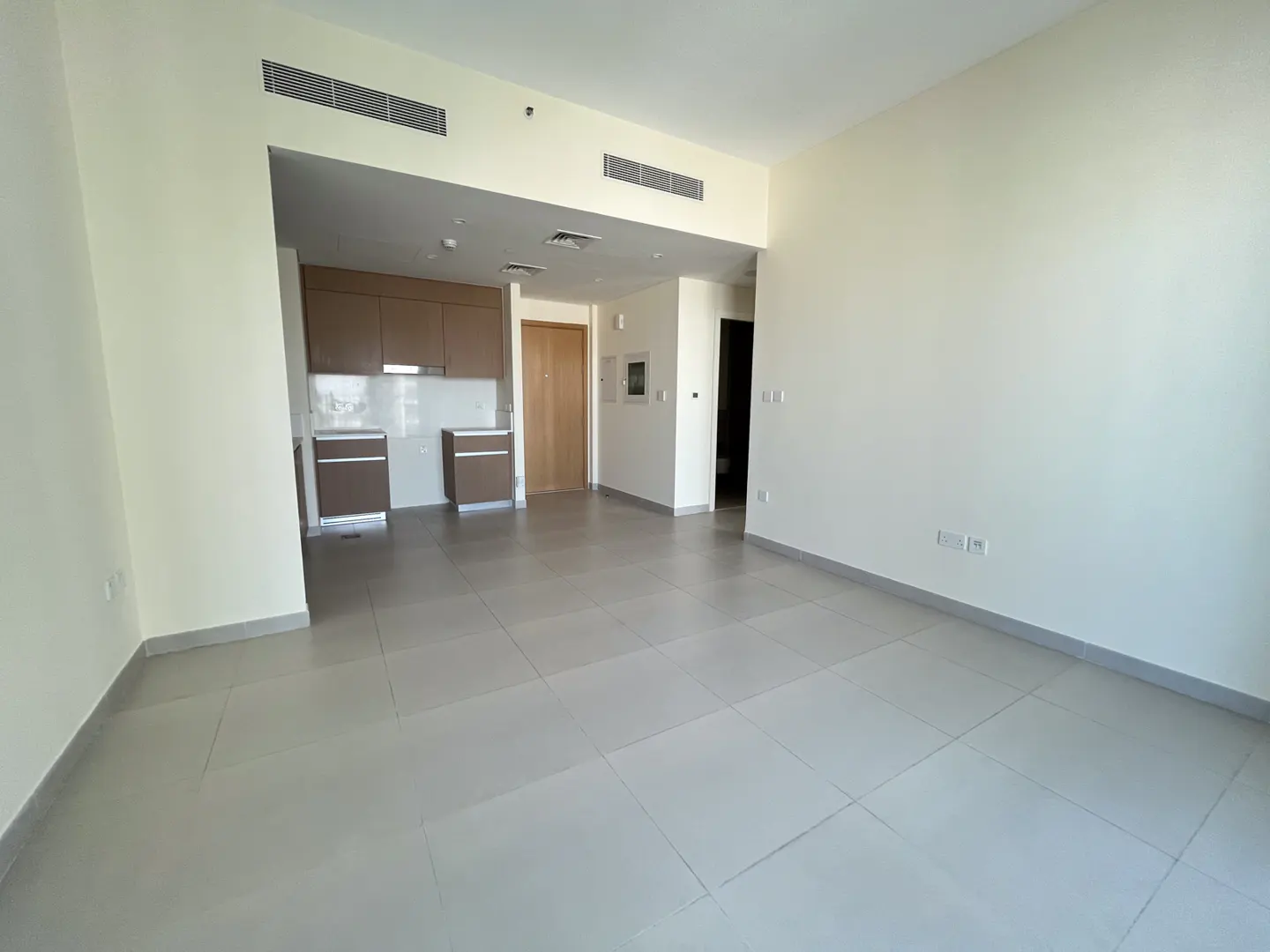 Empty living area with light tile floors, white walls, and an open-plan kitchen with brown cabinets and a wooden door.