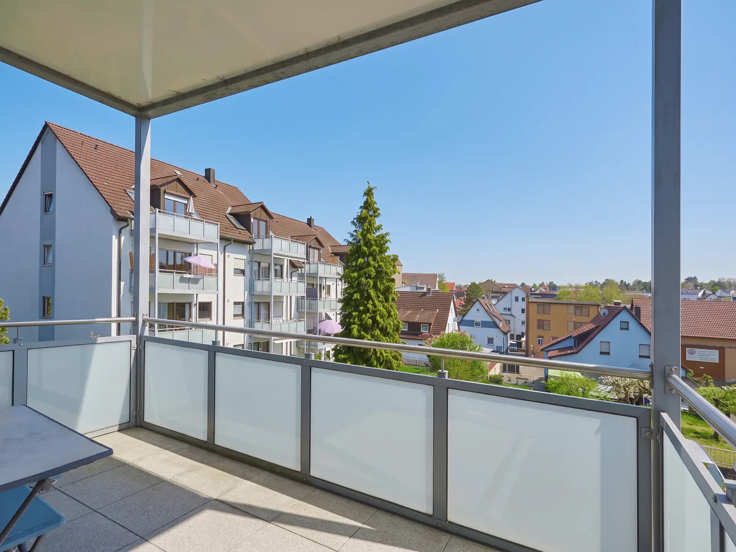 View from a balcony with frosted glass panels and gray metal frames, overlooking a neighborhood with houses and a blue sky.