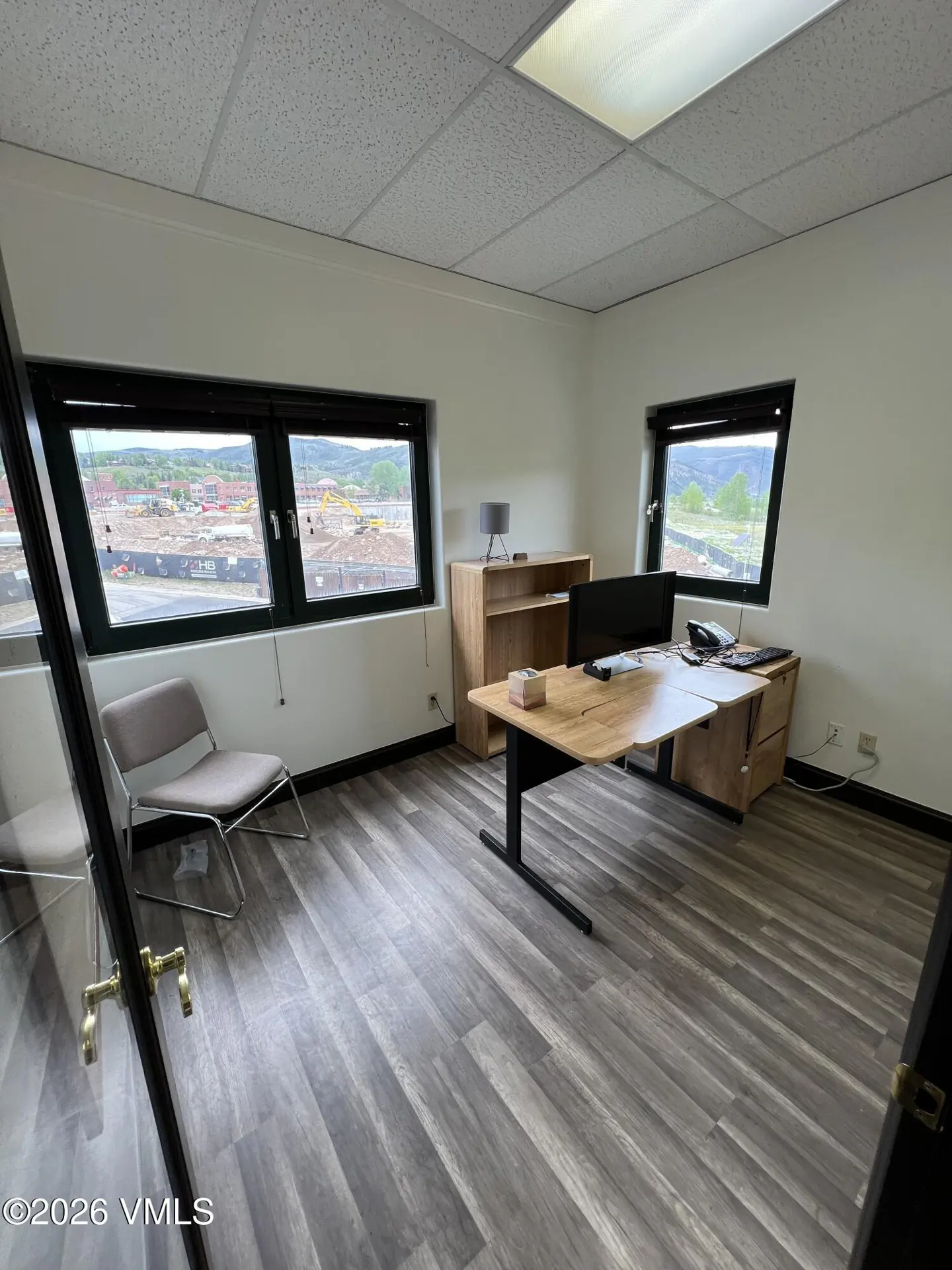 Modern office with wood-look floors, a desk, computer, chair, and large windows overlooking a construction site.