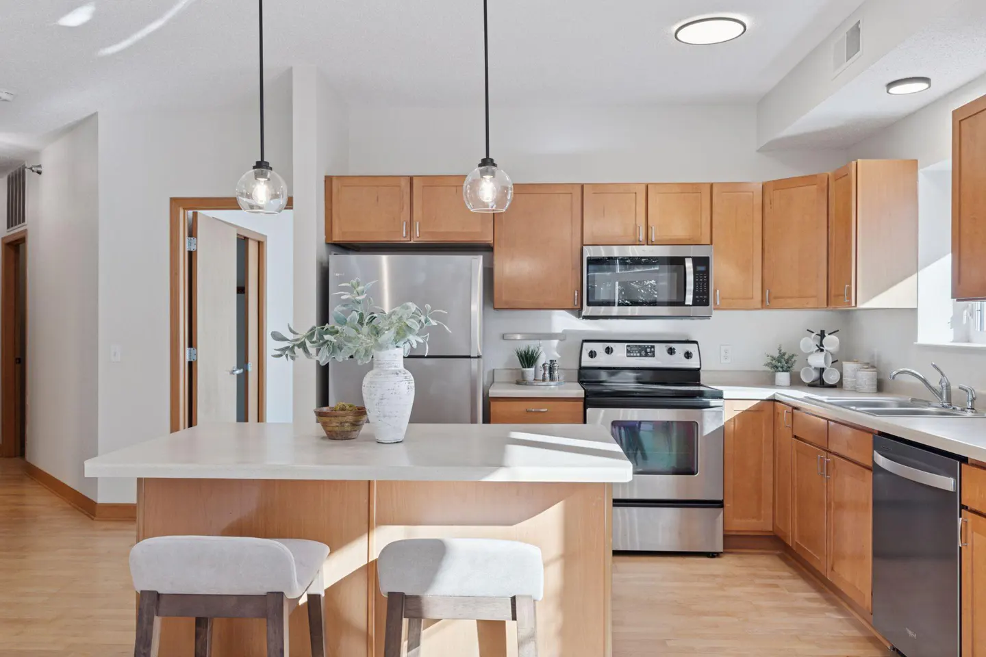 Bright kitchen with wood cabinets, stainless steel appliances, and a white countertop island with two stools. Two pendant lights hang above.
