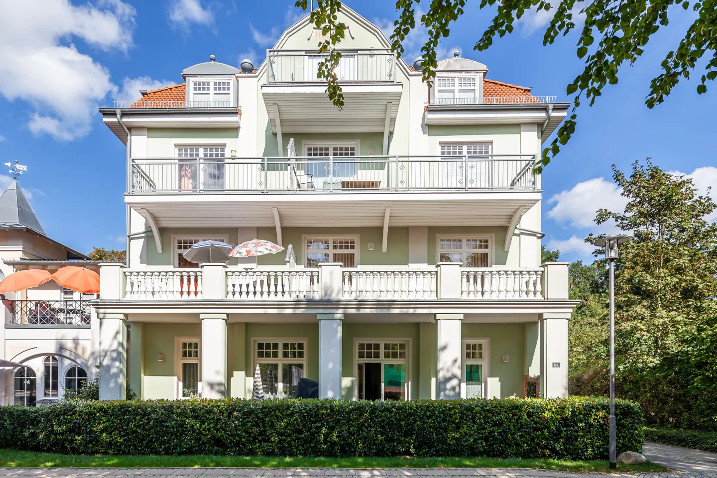 Three-story light green building with white balconies and a red tile roof, framed by green trees and a blue sky.