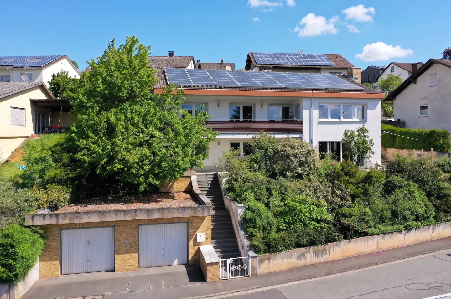 Two-story white house with solar panels on the roof, a brick garage, and a stairway leading to the front door.