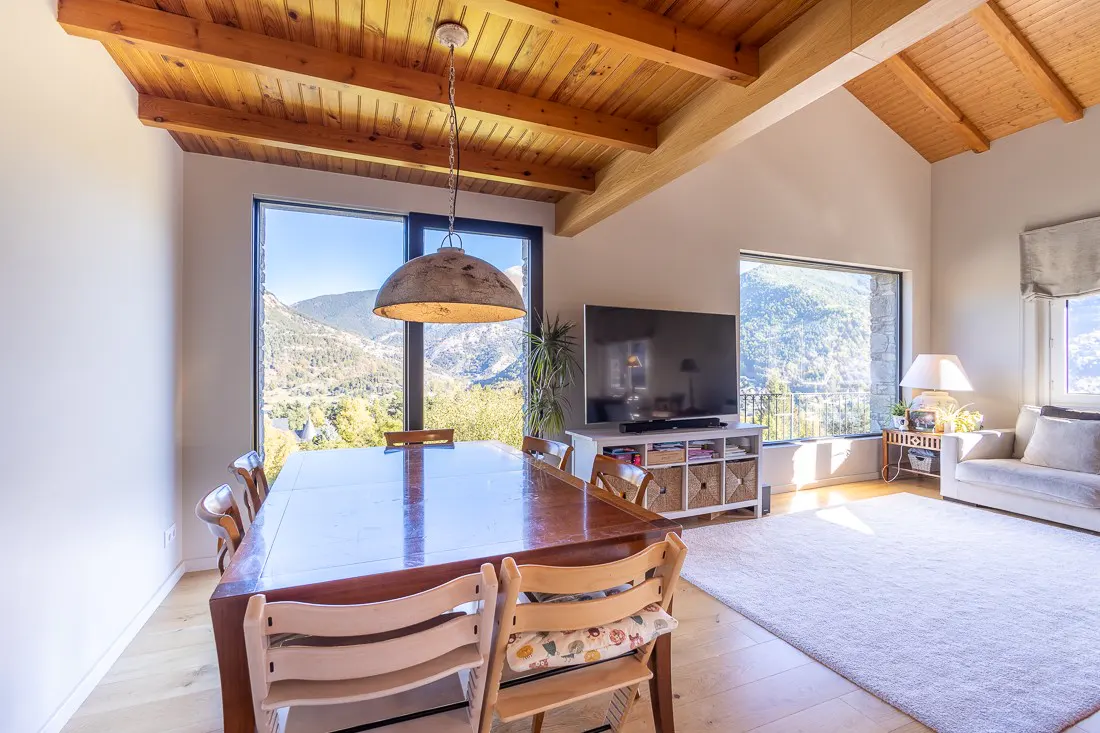 Bright, open-plan living and dining area with wood-beamed ceiling, large windows with mountain views, and a long wooden dining table.