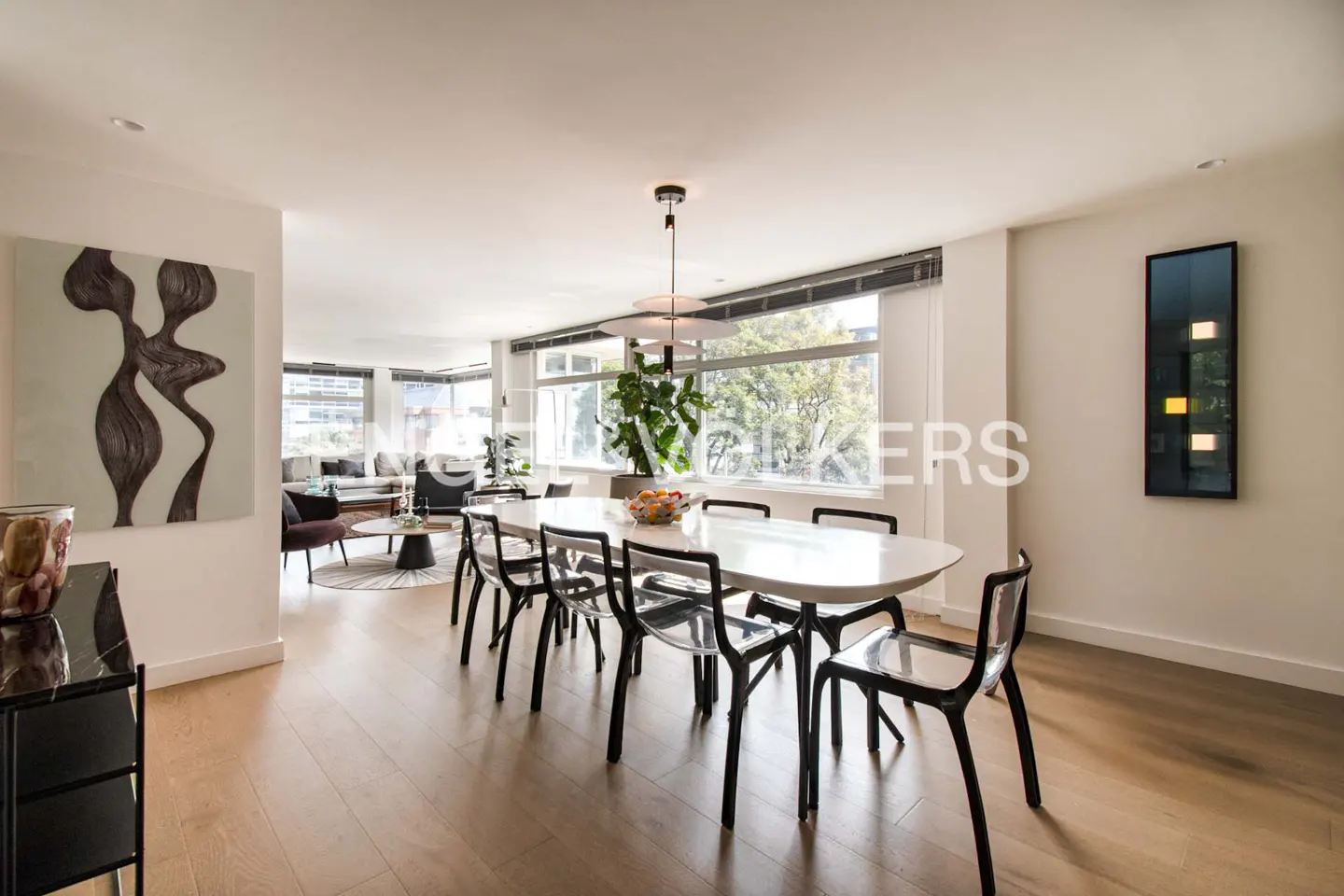 Bright, modern dining room with a white table, black chairs, and wood floors. A living room is visible in the background.