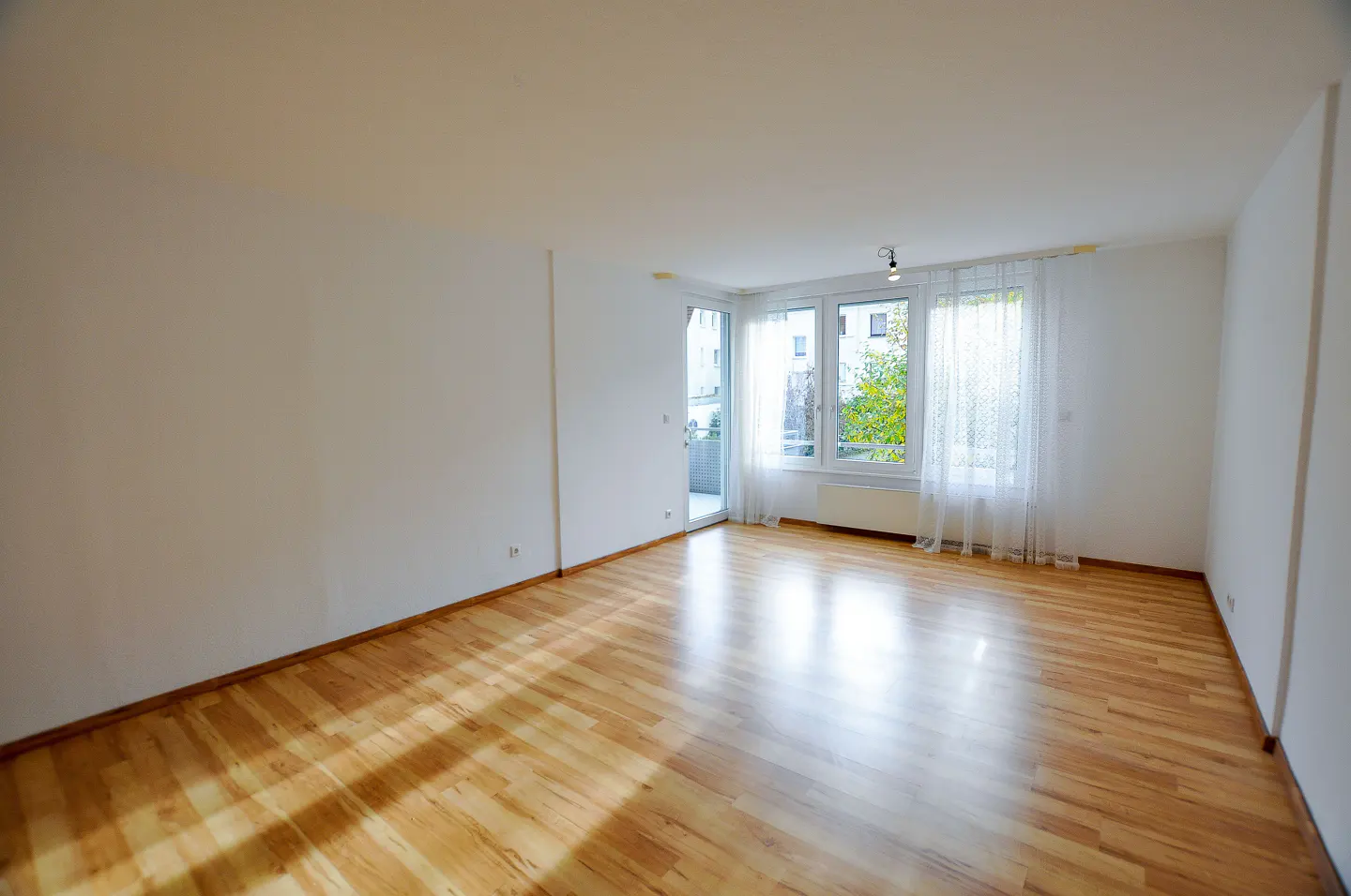 Empty room with light wood floors, white walls, and large windows with sheer curtains. A balcony is visible through a glass door.