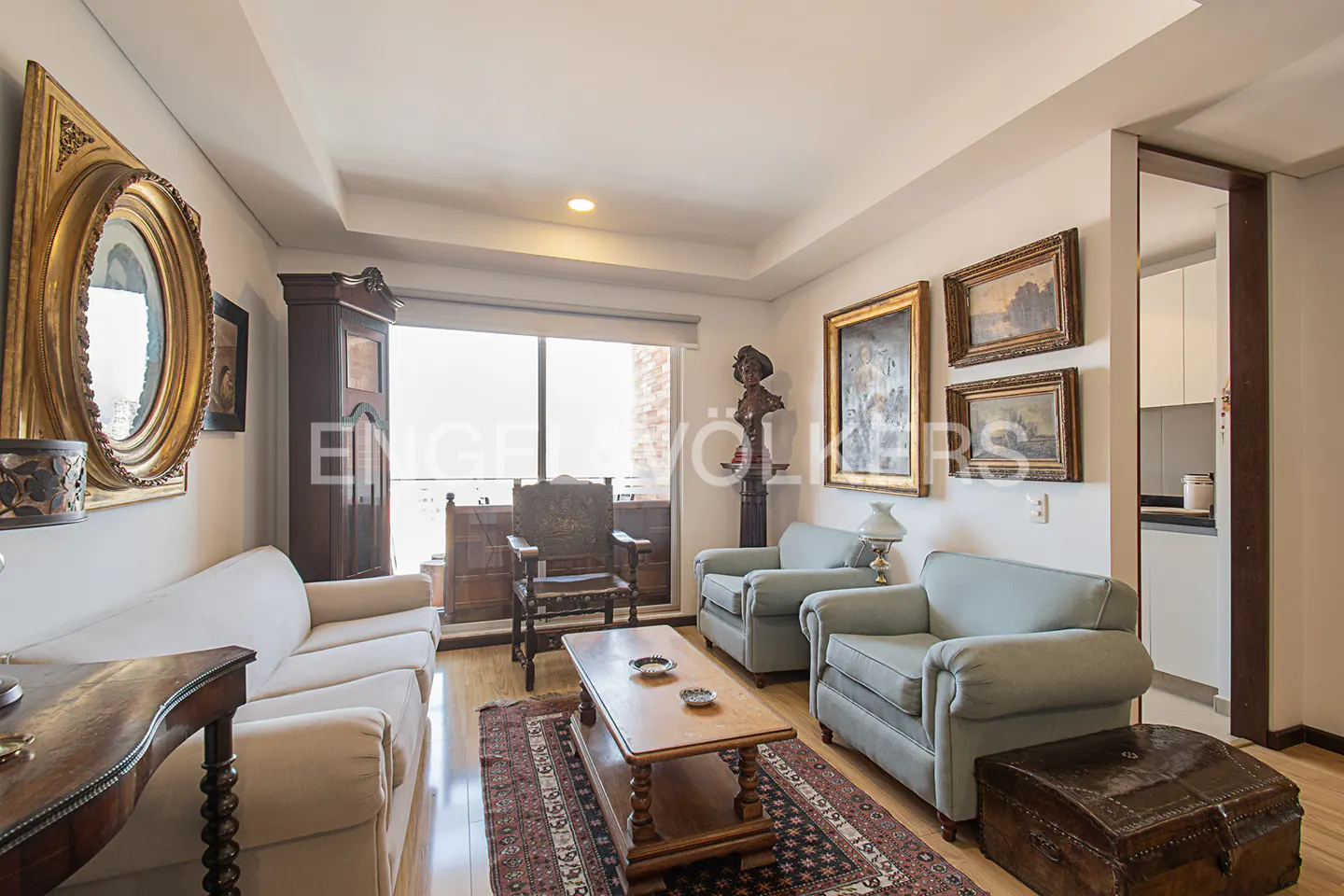 Living room with light blue armchairs, white sofa, wooden table, and antique decor. Large window with balcony view.