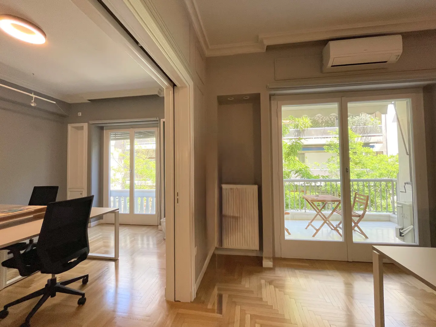 Office space with herringbone wood floors, a desk, and a black chair. A balcony with a table and chairs is visible through sliding glass doors.