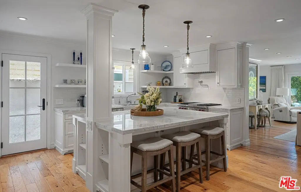 Bright, modern kitchen with white cabinets, marble island with stools, and hardwood floors. Pendant lights hang above the island.