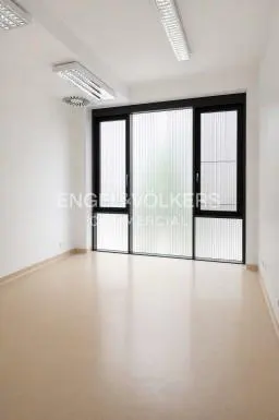 Empty office space with white walls, light flooring, and large windows with black frames and vertical blinds.