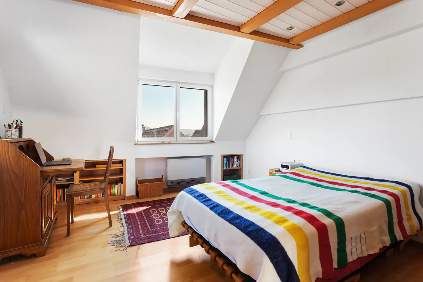Attic bedroom with white walls, wood floors, and exposed beams. A bed with a striped blanket sits near a desk and window.