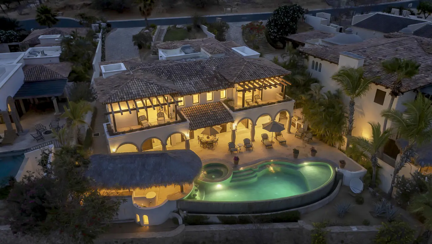Aerial view of a two-story white stucco house with a red tile roof, a pool, and a thatched-roof bar area.