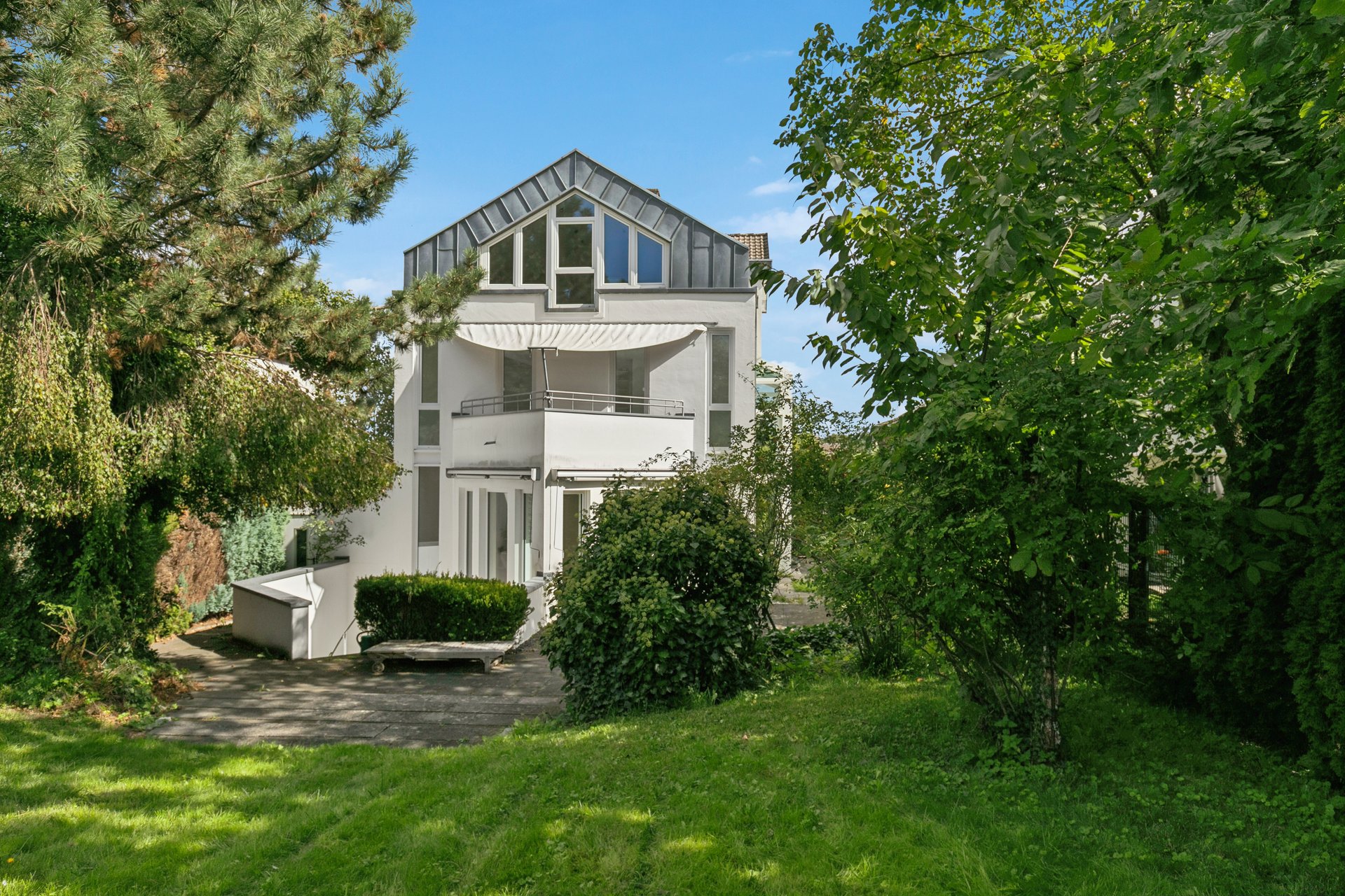 White modern house with a balcony and attic windows, surrounded by lush green trees and a lawn on a sunny day. [AI-generated text]