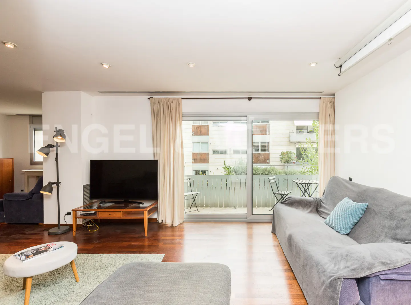 Modern living room with a grey sofa, TV on a wooden stand, floor lamp, and sliding glass doors leading to a balcony.