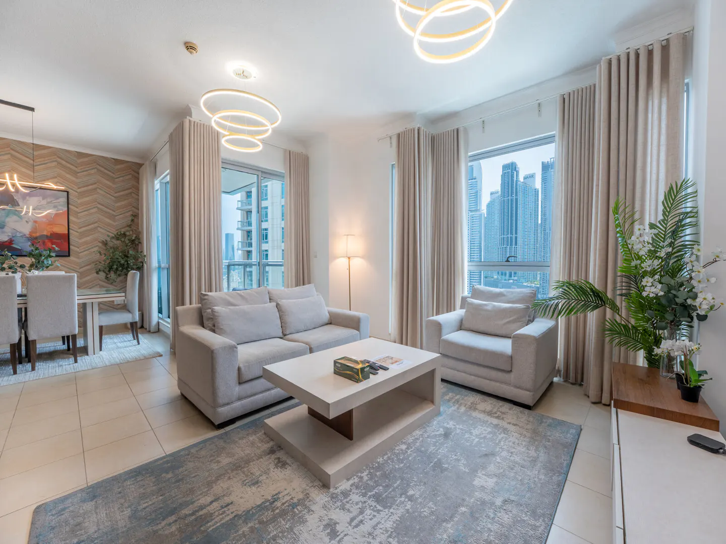 Bright living room with beige sofa, chair, and square coffee table on a blue rug. Large windows show a city view. Dining area visible.