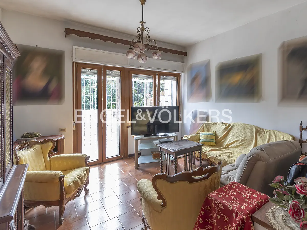 Living room with yellow armchairs, a sofa with a yellow cover, and a TV. French doors lead to a garden. Engel & Völkers logo overlay.