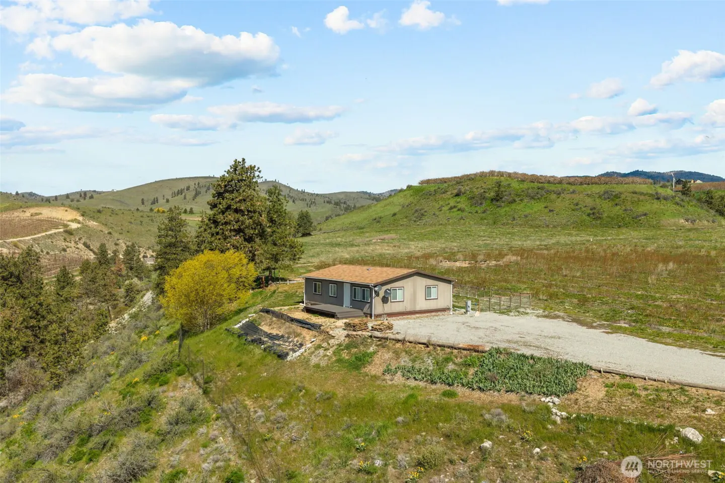 A tan single-story home with a brown roof sits on a grassy hill under a blue sky with scattered clouds.