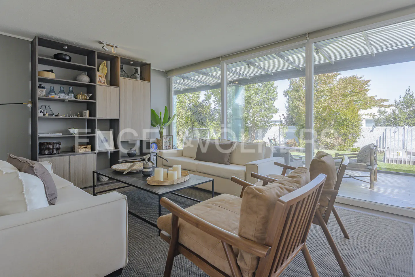 Bright living room with white sofas, wood chairs, and a black coffee table with candles. Large windows overlook a green yard.