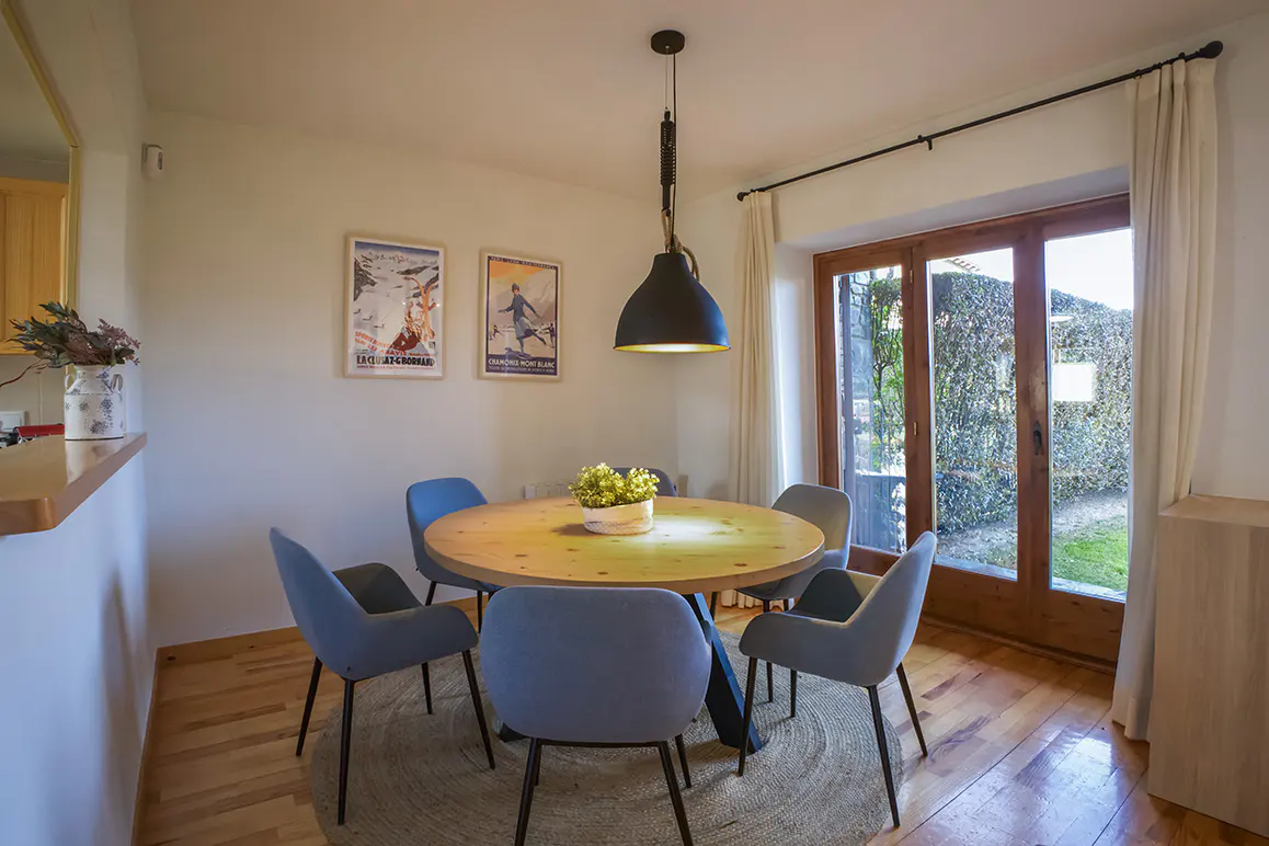 A dining room with a round wooden table, six blue chairs, and a black pendant light. Two framed posters hang on the wall. A door leads to a garden.