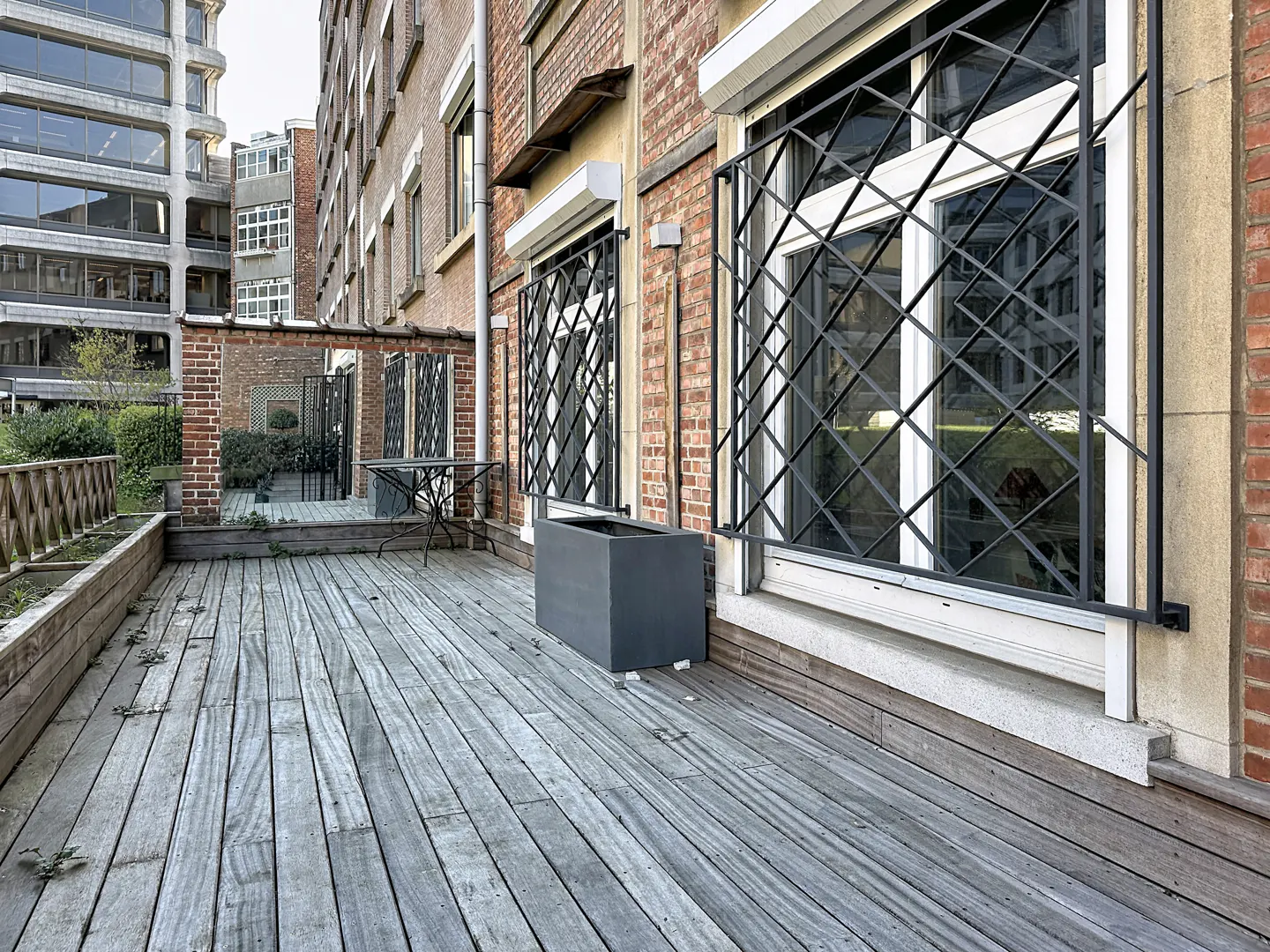 Exterior view of a wooden deck with a brick building, windows with black iron bars, and a gray planter.