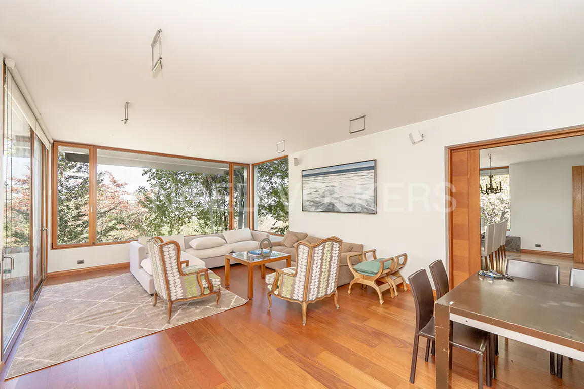 Bright living room with wood floors, large windows, and a view of trees. A white sectional sofa and patterned armchairs sit on a geometric rug. A dining table is visible in the foreground.