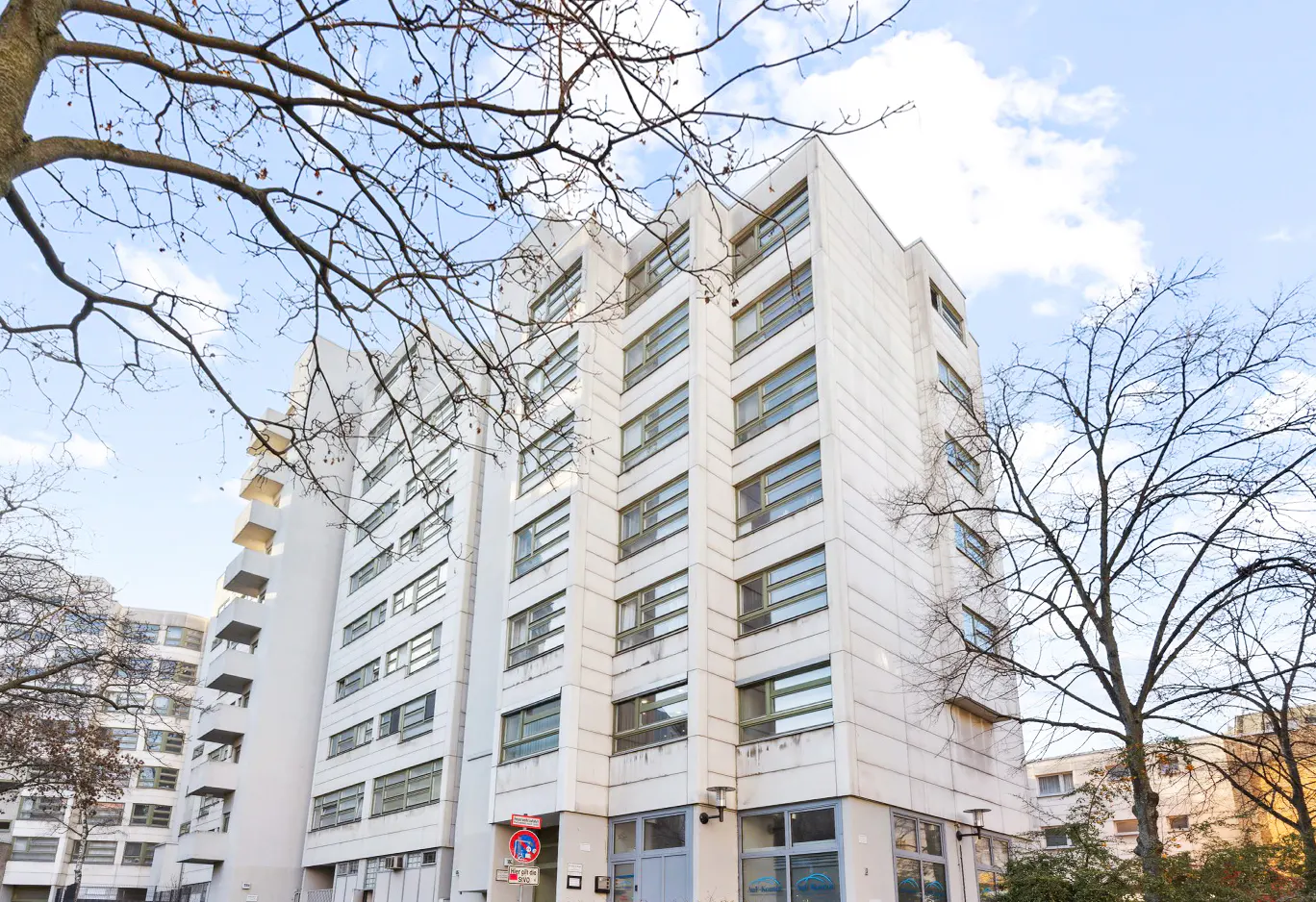 Exterior view of a tall, white apartment building with many windows against a blue sky with bare trees.