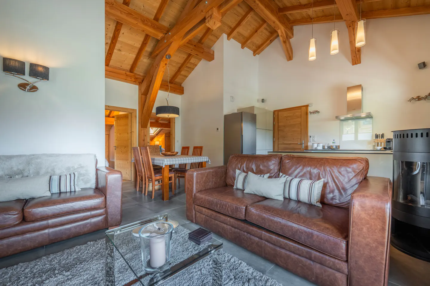 Living room with brown leather sofas, glass table, dining table, and kitchen in the background. The ceiling has wooden beams.