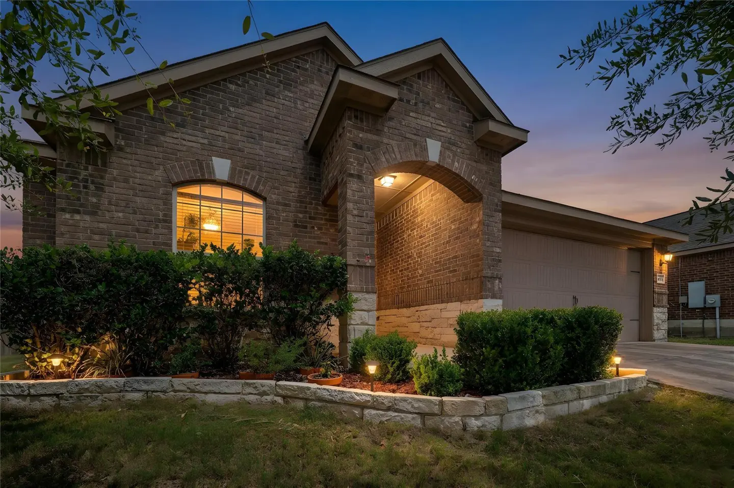 Brick house at dusk with an arched entryway, a two-car garage, and manicured landscaping with stone edging.