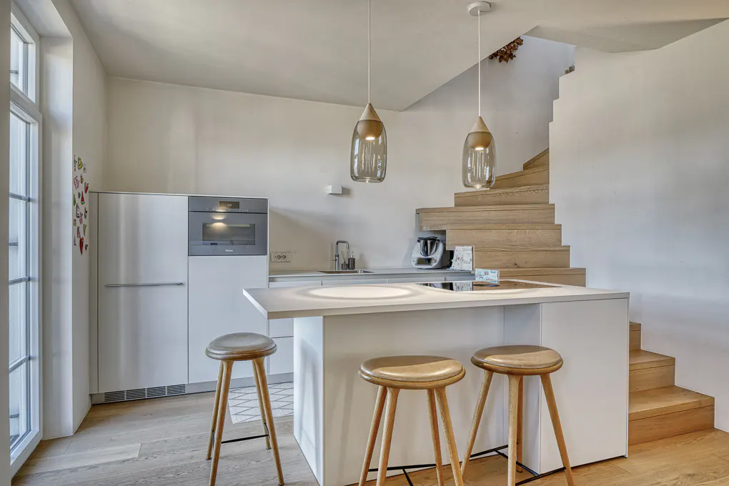 Bright kitchen with white cabinets, island with three wooden stools, and wood stairs leading up. Two pendant lights hang above the island.