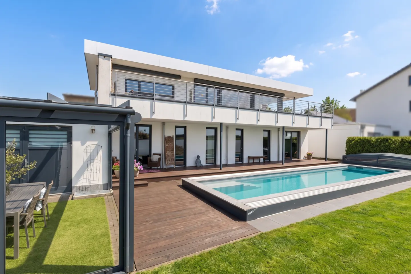 Modern white two-story house with a balcony, wood deck, and a rectangular swimming pool on a sunny day.