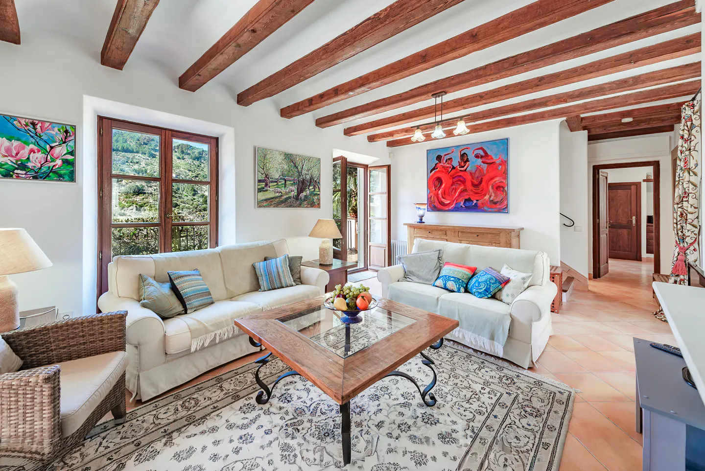 Bright living room with white sofas, wood beams, and a patterned rug. Art adorns the walls, and a window offers a view of lush greenery.