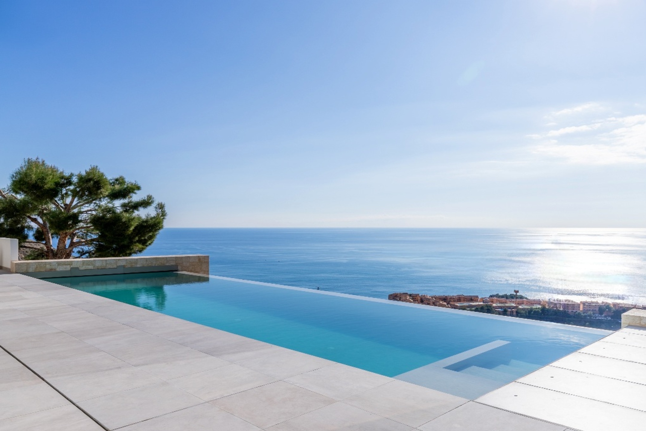 Infinity pool with turquoise water overlooking the blue ocean and a coastal town under a clear sunny sky. [AI-generated text]