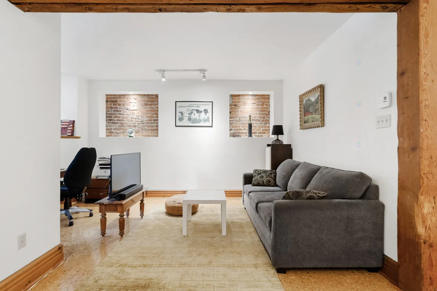 Living room with white walls, exposed brick alcoves, and a gray sofa. A desk and chair are visible in the background.