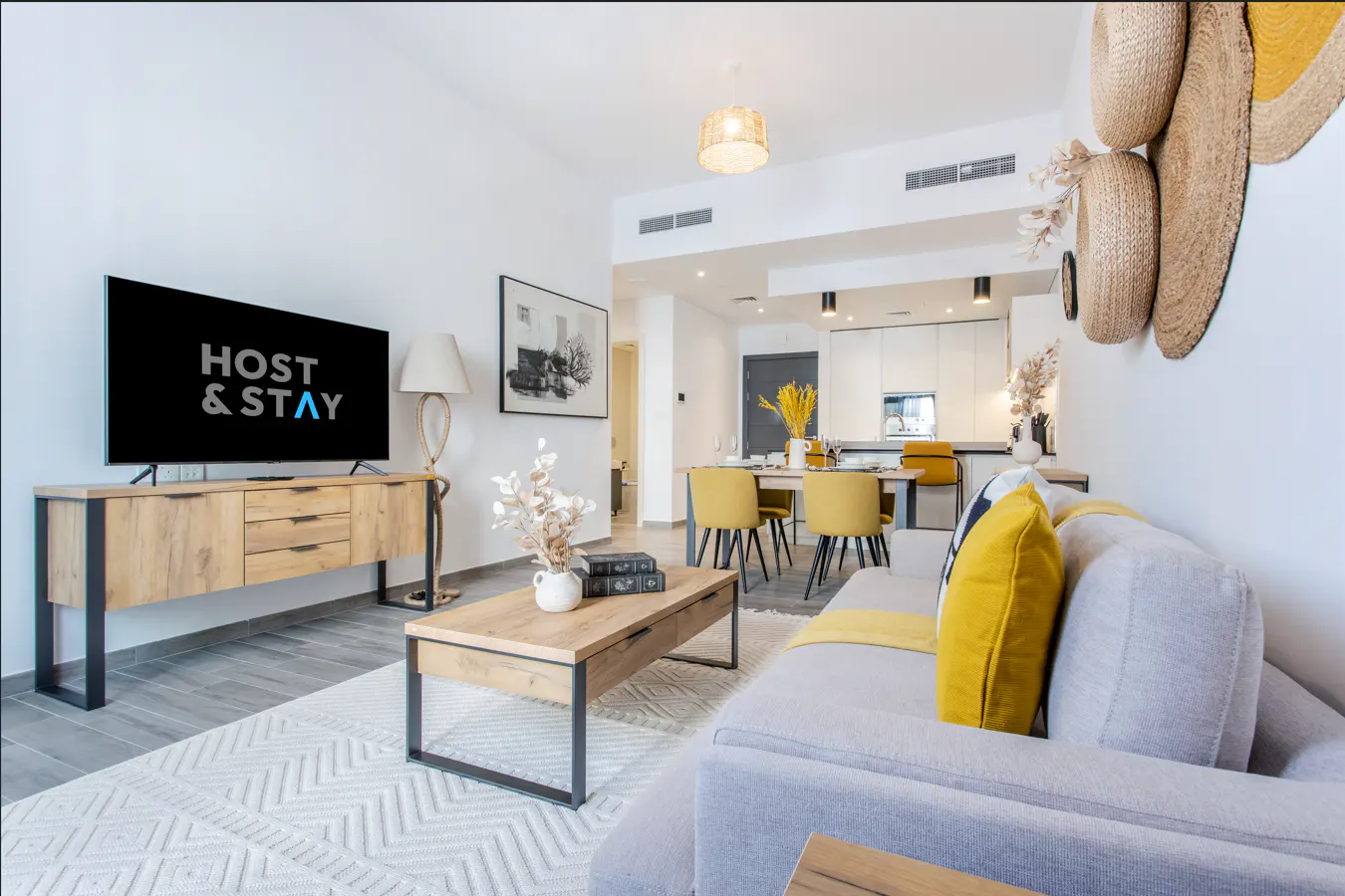 Bright, modern living room with a TV displaying "Host & Stay", a gray sofa with yellow accents, and an open-plan kitchen and dining area.