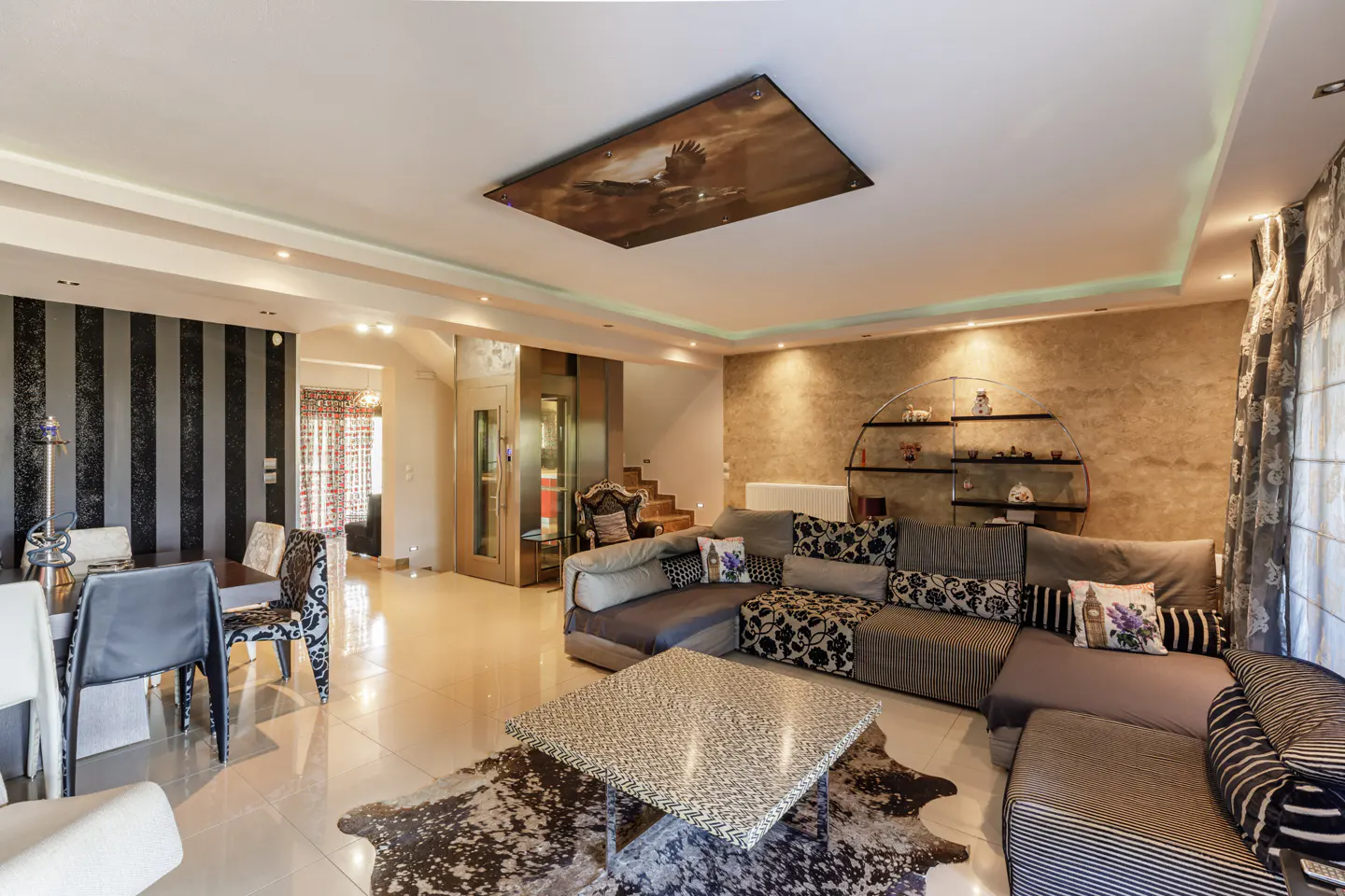 A modern living room with a large gray sectional sofa, a cowhide rug, and a glass-topped coffee table. A dining area is visible in the background.