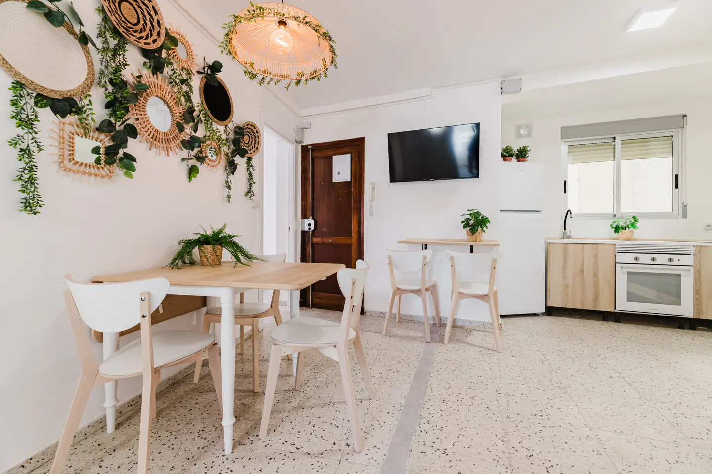 Bright, modern apartment interior with a dining table, white chairs, and a kitchen area with light wood cabinets. Woven wall decorations and plants add a natural touch.