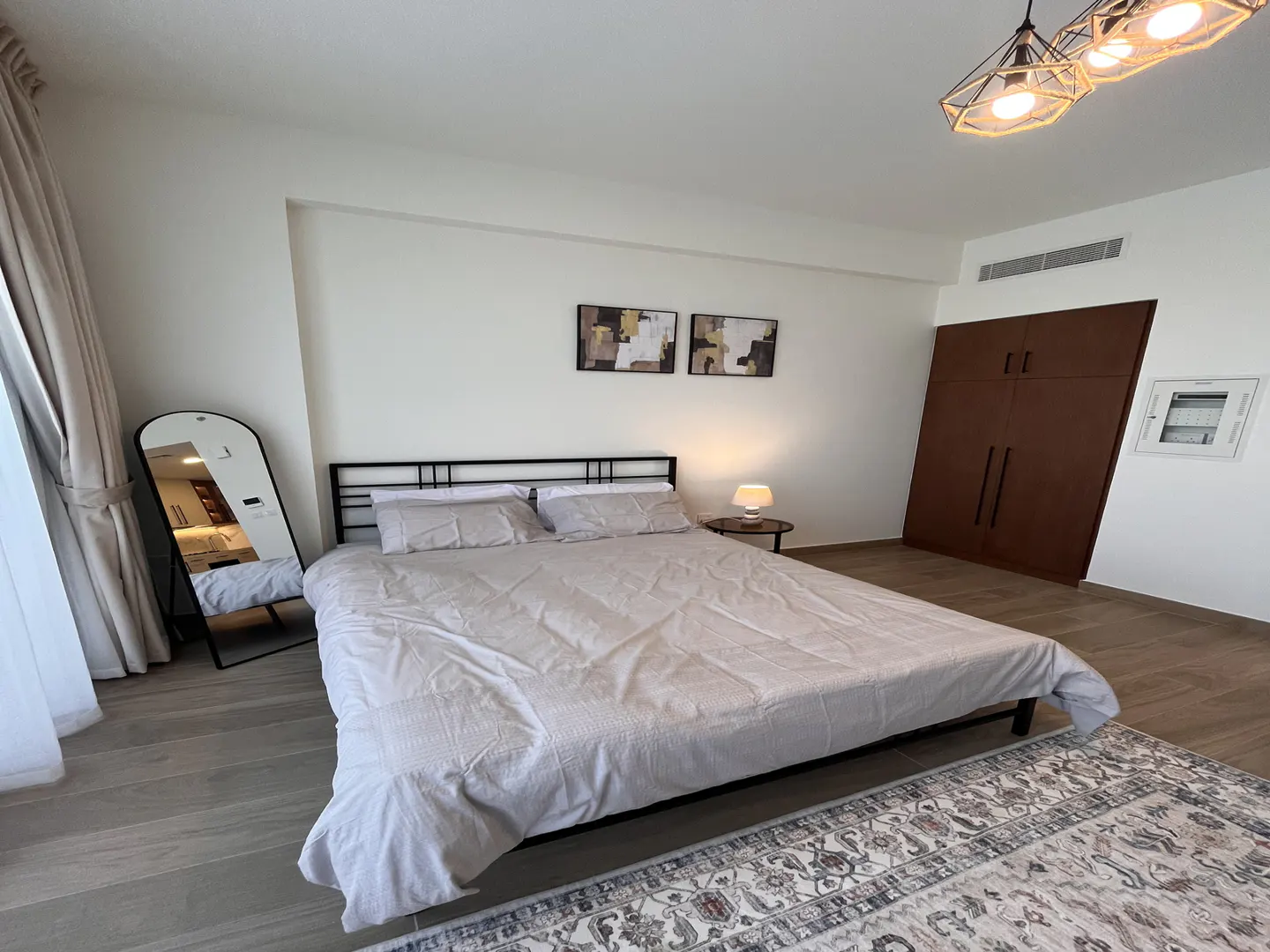 Bedroom with a black metal bed frame, light gray bedding, and a full-length mirror. A patterned rug is on the floor, and modern art hangs above the bed.