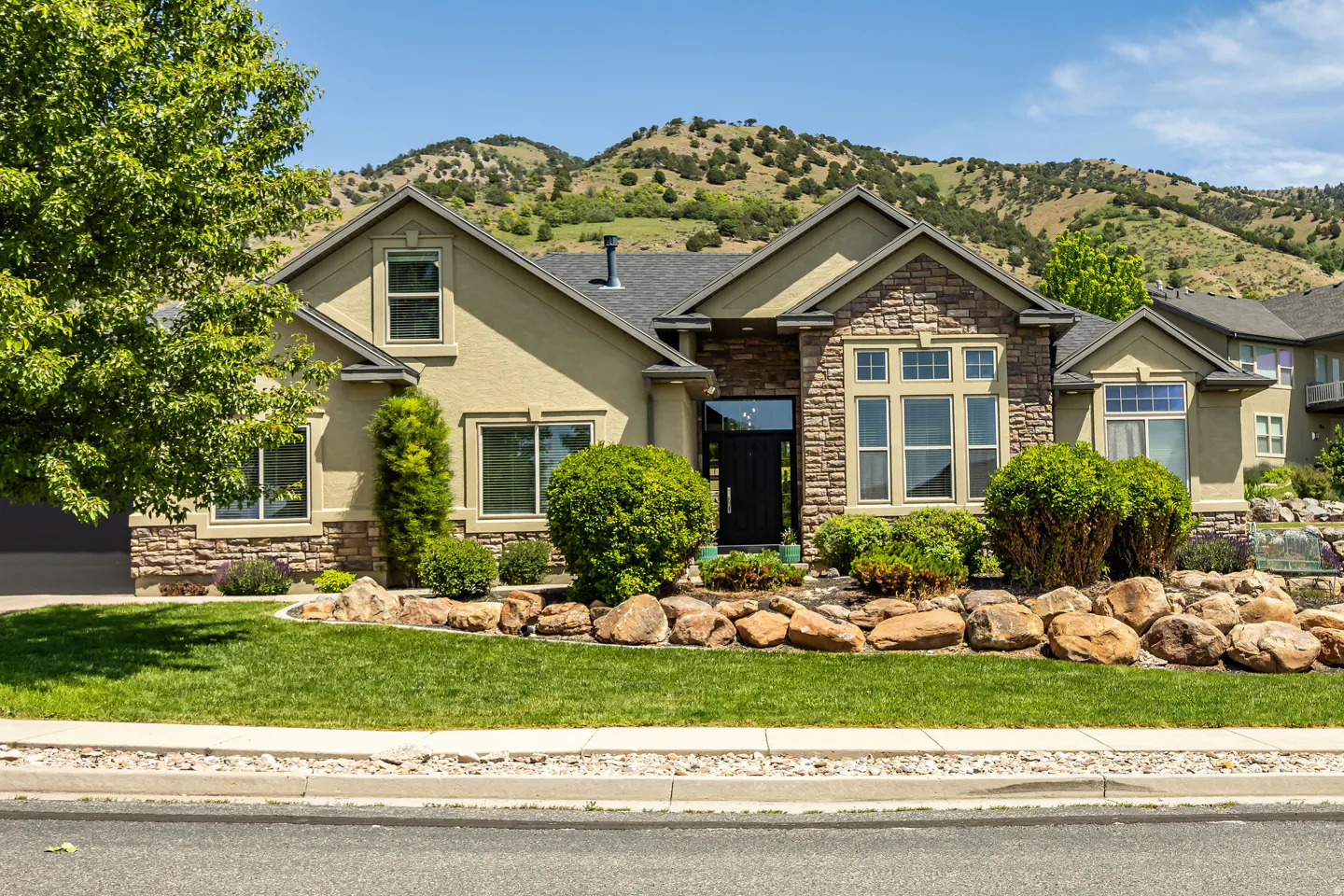 Tan house with stone accents, dark front door, manicured lawn, and large boulders against a backdrop of rolling hills.