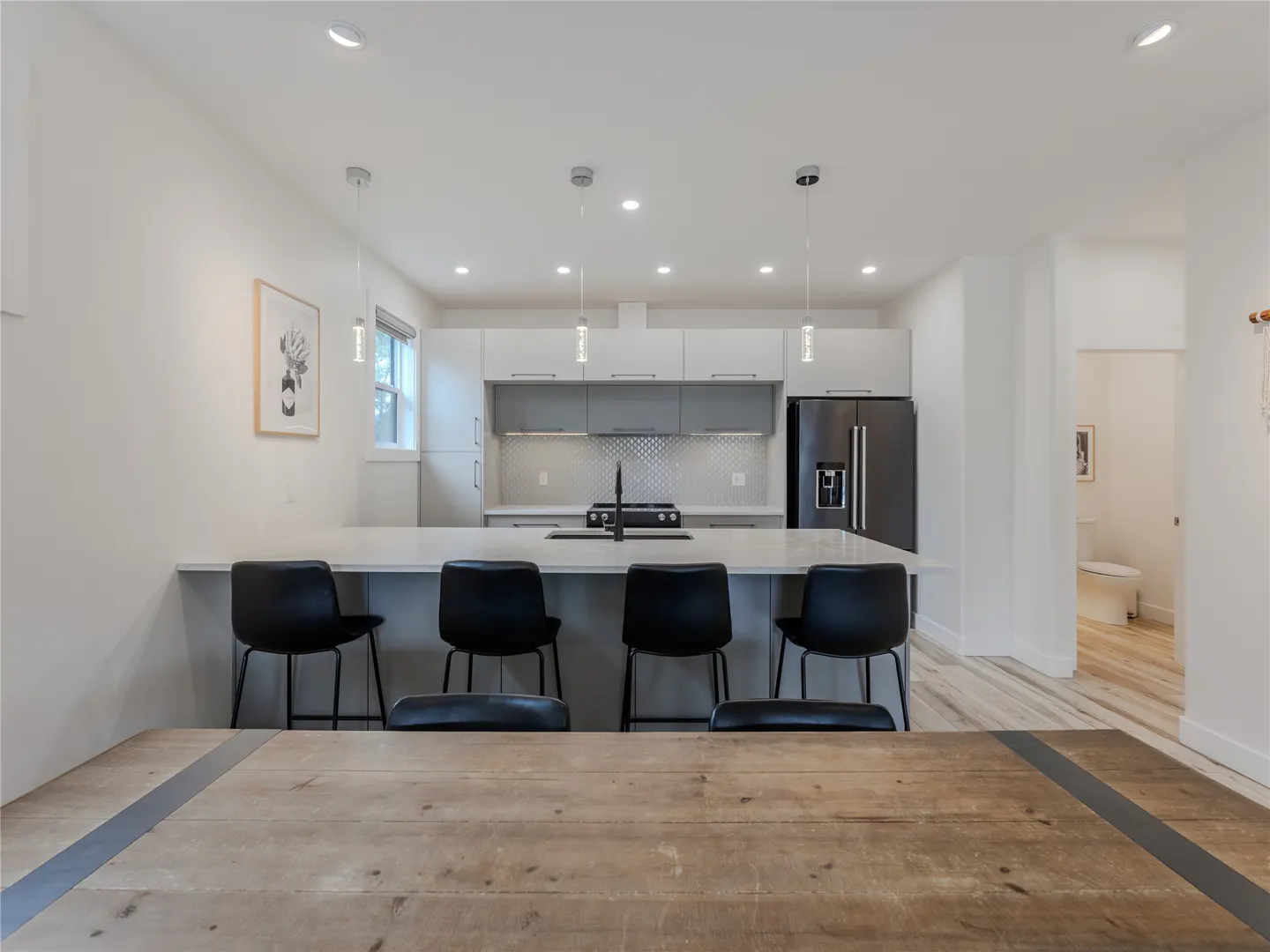Modern kitchen with white cabinets, a large island with four black stools, and a rustic wooden dining table in foreground.