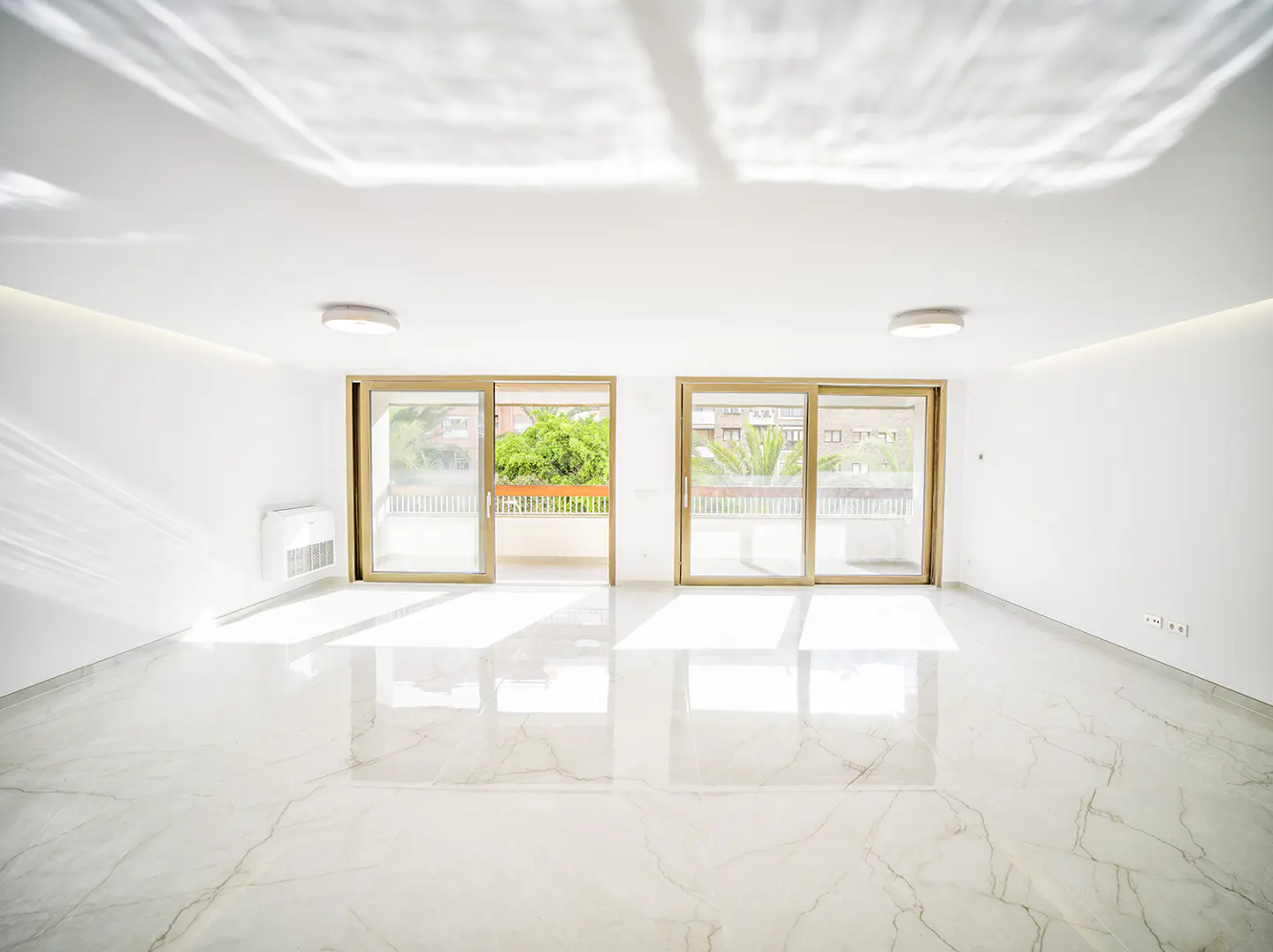 Bright, empty room with white walls, marble floors, and two sliding glass doors leading to balconies with green trees visible.