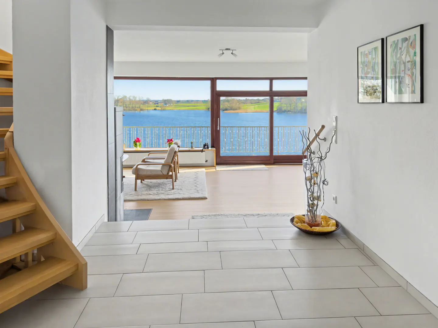 Interior view of a modern home with a wooden staircase, tiled floor, and a living room with a lake view through large windows.